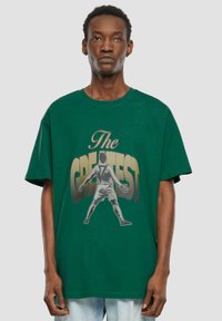 Grøn bomulds t-shirt med en afslappet pasform, prydet med et centralt grafisk motiv af en fodboldspiller og teksten "The GREATEST" i fed gul.