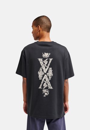 Homme aux cheveux courts portant un T-shirt noir ample avec des lettres verticales blanches ornées "VXA" dans le dos sur un fond uni.