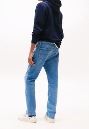 Person steht, trägt hellblaue Jeans, marineblauen Hoodie und weiße Turnschuhe mit blauen Details, vor einem schlichten weißen Hintergrund.