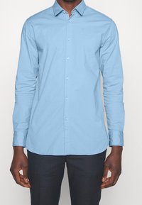 Ljusblå skjorta med knäppning i slätt bomullstyg, med spetsig krage och lång ärm med knäppta manschetter.