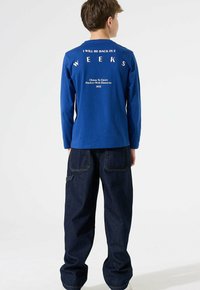 Junge steht mit dem Rücken zur Kamera und trägt ein blaues langärmliges Hemd mit dem Text "Ich bin in 2 Wochen zurück" und lockere, dunkle Jeans.