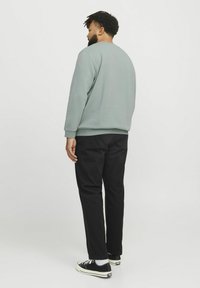Lichtgroene sweatshirt met een ronde hals en geribbelde manchetten, gecombineerd met zwarte broek. De persoon draagt zwarte sneakers met witte zolen.