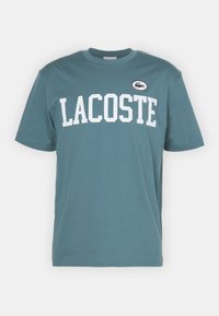 Tyrkysové bavlněné tričko s velkým bílým nápisem "LACOSTE" a kulatou nášivkou s krokodýlem na hrudi. Krátké rukávy, kulatý výstřih.