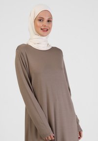 Braunes, langärmliges Kleid aus weichem Stoff mit lockerer Passform. Ein gewickelter, hellfarbener Hijab ergänzt das Gesamtoutfit.