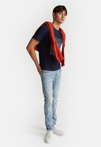 Námořnické tričko s barevným potiskem, světle modré džíny a bílé tenisky. Červený svetr přehozený přes ramena.