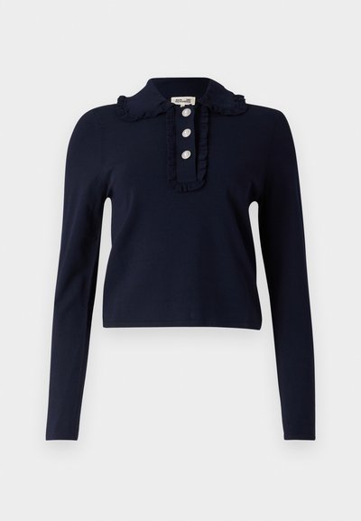 Jūrų mėlynos spalvos ilgomis rankovėmis viršus su raukiniais apykakle, trimis dekoratyviniais mygtukais prie kaklo ir prigludusia forma. Minkšta medžiaga ir sutrumpintas ilgis.