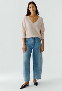 Hellrosa V-Ausschnitt-Pullover mit gerippter Textur, kombiniert mit locker sitzenden blauen Jeans. Frau trägt schwarze spitze Ballerinas und steht vor einem schlichten Hintergrund.