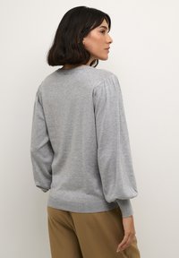 Pull en gris avec des manches bouffantes et des poignets côtelés. Tissu tricot doux. Encolure arrondie. Porté sur un pantalon beige clair.