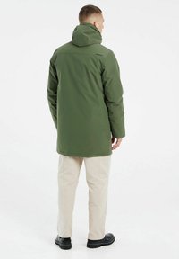 Parka verte imperméable avec capuche, présentant une coupe droite et un logo dans le dos, associée à un pantalon clair à coupe décontractée.