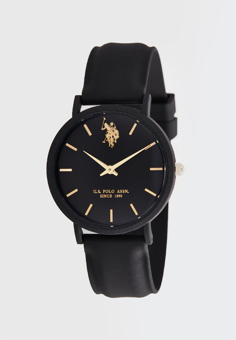 Reloj de pulsera de silicona negra con una caja redonda, acentos dorados, esfera minimalista y logotipo. Presenta marcadores horarios de tipo palo y manecillas delgadas.