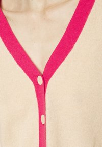Beige neulecardigan, jossa on V-päänti, ja kirkkaan vaaleanpunainen röyhelökaulus sekä kolme vaaleancreme väriä olevaa nappia etuosassa. Pehmeä rakenne.