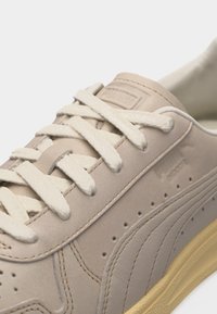 Μπεζ δερμάτινο αθλητικό παπούτσι με εκρού κορδόνια, ραφές, διάτρητα σχέδια και κίτρινη λαστιχένια σόλα, εμφανιζόμενο σε κοντινή όψη.