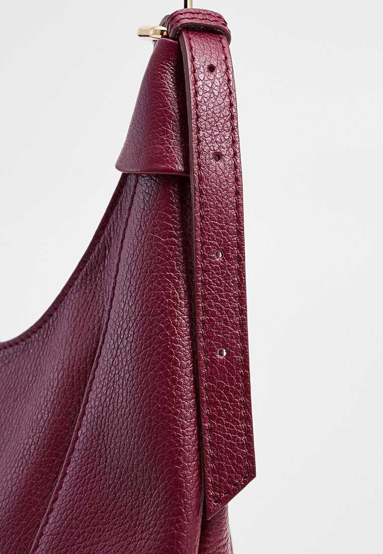 Sac à main en cuir bordeaux avec une finition texturée, présentant un corps plissé large et une bandoulière réglable avec des coutures décoratives. Accents en or.