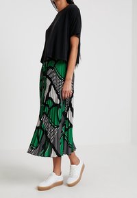 Černý cropped top v kombinaci s dlouhou zelenou sukní s vzorem, která obsahuje černé a bílé geometrické vzory, doplněný bílými teniskami.