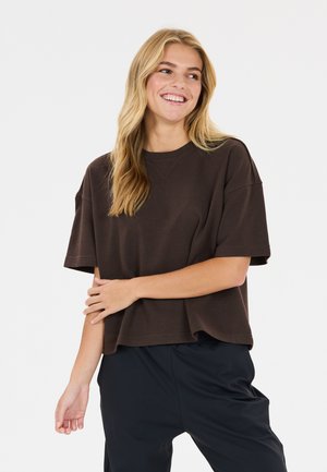 Ung kvinde smiler iført en løs mørkebrun kortærmet skjorte og sorte bukser mod en ensartet lys baggrund.