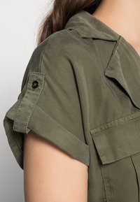 Camicetta verde oliva con maniche arrotolate, tasca frontale e texture liscia. Presenta un colletto strutturato e accenti di cucitura.