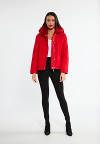 Rode pufferjas met een nepbont capuchon, rits- en drukknoopsluiting, en lange mouwen. Gecombineerd met zwarte leggings en enkellaarzen.