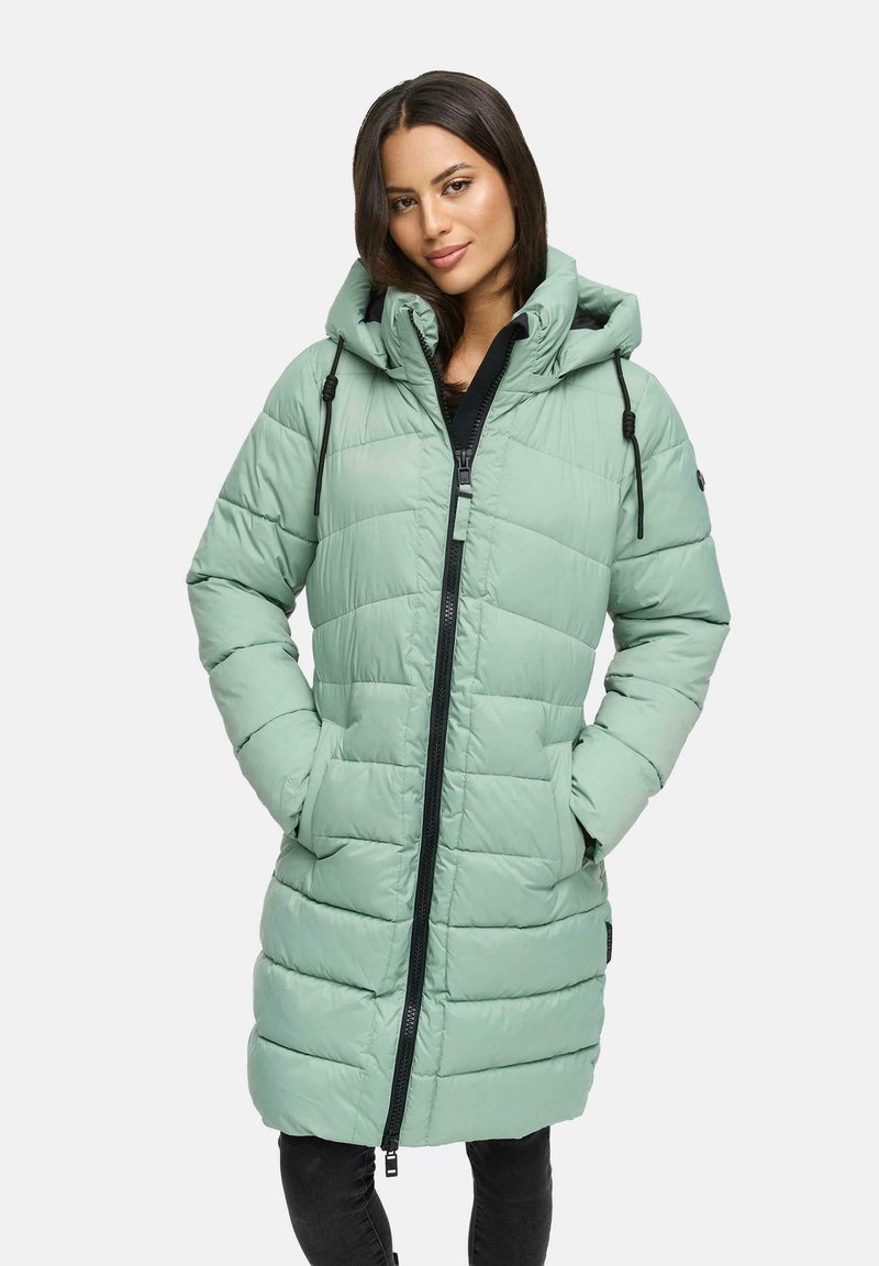 Kvinna klädd i en ljusgrön vadlång vadderad jacka med huva och svart dragkedja, med händerna i fickorna, stående mot en enfärgad bakgrund.