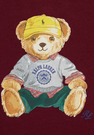 Ralph Lauren meškiukas su grafika, vilkintis pilku megztiniu su mėlynu logotipu, švarkeliu su languotais apykakle, geltonu kepuraitė ir žaliais šortais ant bordo fono.