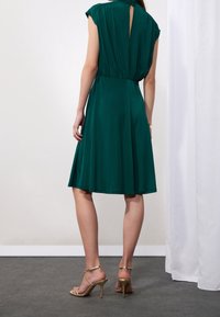 Kvinna som står i en smaragdgrön knälång klänning med samlad midja och guldiga högklackade sandaler, visad från baksidan mot en vit vägg.