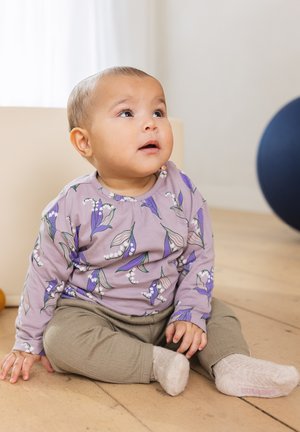 Langärmliges lila Hemd mit Blumenmuster, beige Hose und blassen Socken; sitzt auf einem Holzboden in einer hellen Innenumgebung.
