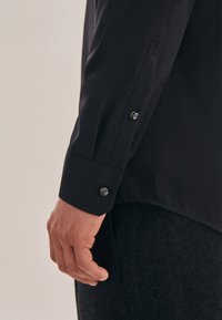Zwart langarmshirt met knoopmanchetten, gemaakt van gladde stof; heeft een subtiele textuur en een getailleerde pasvorm, waarbij een deel van een hand zichtbaar is.