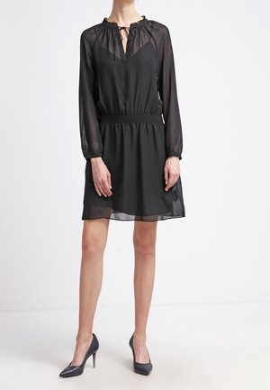 Kobieta ubrana w czarną, półprzezroczystą sukienkę z długim rękawem sięgającą do kolan, z taliowanym pasem i czarnymi szpilkami, stojąca na białym tle.