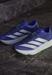 Violetti juoksukengät, joissa on valkoisia raitoja, verkkopinta ja pehmeä pohja, jossa on vihreä yksityiskohta kannassa. Teksturoitu suunnitteluteksti on läsnä.