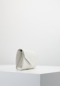 Bolso de mano de satén blanco con tapa triangular y cadena plateada descansando sobre un pedestal blanco contra un fondo liso.
