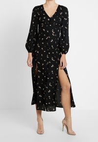 Sort blomsterkjole med lange pufærmer, V-hals, knaplukning, sideslids og et svømmede design. Har pink rosemønstre gennem hele.