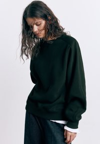 Žena valovite smeđe kose u širokoj crnoj dukserici i tamnim hlačama, gleda dolje, s vidljivim bijelim rukavom košulje.