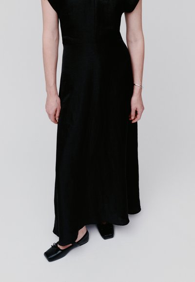 Άτομο που φοράει αμάνικο μαύρο φόρεμα με ασημένιο βραχιόλι στον δεξί καρπό, στέκεται με σταυρωμένα πόδια, φορώντας μαύρα παπούτσια με τετράγωνη μύτη.