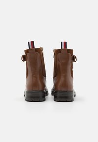 Botas de piel marrón hasta el tobillo con cierre de cremallera, punta redonda y suela de goma negra. Presenta acentos textil en contraste en el talón.