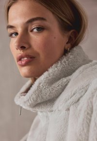 Grijze jas met hoge kraag van nepbont met een zachte textuur, voorzien van een zilverkleurige rits aan de voorkant. Close-up weergave met nadruk op gelaatstrekken.