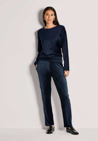 Nachtblaue Samt-Loungewear mit langärmeligem Oberteil in dezentem Leopardenmuster und passender Hose, kombiniert mit schwarzen Loafers.