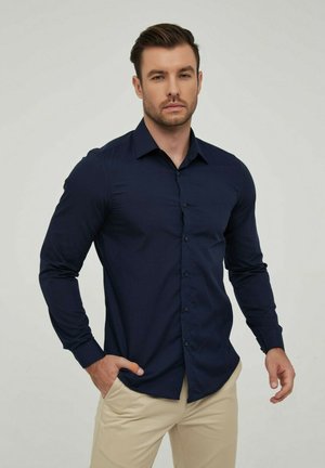 Chemise à manches longues navy avec fermeture à boutons, col classique, design ajusté, en tissu lisse, associée à un pantalon beige clair.