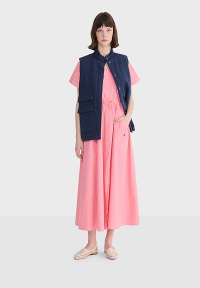 Femme portant une robe midi rose à boutons sur le devant et un gilet sans manches bleu marine avec des poches, debout avec une main dans la poche de la robe.