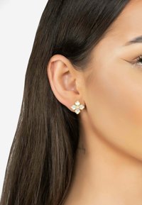 Gold flower-shaped earrings with four white pearl-like stones and a central crystal, set against a smooth, light skin background.