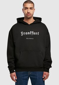Felpa con cappuccio nera realizzata in tessuto morbido, con una grande stampa grafica che dice "Frankfurt" in caratteri bianchi stilizzati e una tasca frontale.
