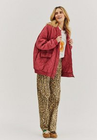 Prešívaná burgundská bunda s rebrovanými manžetami, skombinovaná s béžovými nohavicami s leopardím vzorom. Oblečenie obsahuje biely grafický tričko pod tým.
