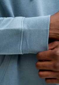 Poignet de sweat-shirt bleu clair avec texture côtelée, montrant des coutures précises. Une main saisit le tissu, mettant en valeur la douceur du matériau.