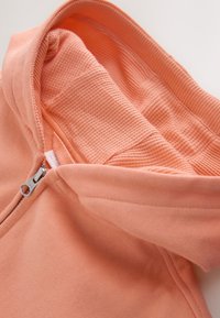 Lähedalt võetud pilt virsikuvärvi kapuutsist, kus on näha metallist tõmblukk ja kapuutsi sees tekstuurse mustriga vooder.