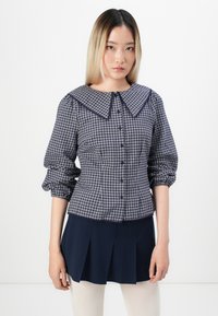 Camicia a quadri blu navy e bianca con un ampio colletto, apertura frontale con bottoni e maniche arricciate. Abbinata a una gonna plissettata blu navy.