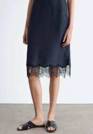 Femme portant une robe bleu marine avec une bordure en dentelle noire à l'ourlet, associée à des sandales à brides croisées noires, debout sur un sol gris.