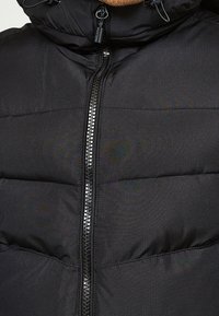 Černá prošívaná bunda s vysokým límcem a předním zipem. Tkanina má jemnou strukturu s horizontálním prošíváním na těle.