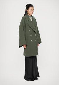 Olive green double-breasted coat with large lapels, oversized sleeves, and silver buttons. Worn over a textured high-neck top and black skirt.