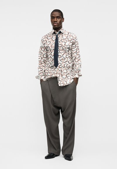 Άνδρας στέκεται με τα χέρια στις τσέπες, φορώντας λευκό πουκάμισο με σχέδια, μαύρη γραβάτα, φαρδιά γκρι παντελόνια και μαύρα παπούτσια, σε απλό φόντο.