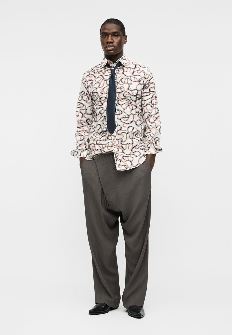 Uomo in piedi con le mani in tasca, indossa una camicia bianca con motivi, cravatta nera, pantaloni grigi larghi e scarpe nere su uno sfondo neutro.