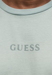 Ανοιχτό γκρι-πράσινο μπλουζάκι με στρογγυλή λαιμόκοψη που δείχνει το αμυδρό λογότυπο "GUESS" τυπωμένο σε ελαφρώς σκουρότερη απόχρωση στο στήθος.