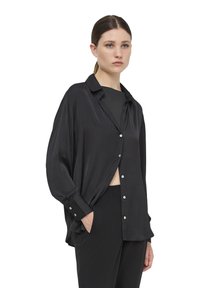 Camicia nera a maniche lunghe in tessuto morbido, con colletto, chiusura a bottoni e vestibilità rilassata. Abbinata a un pantalone slim nero coordinato.